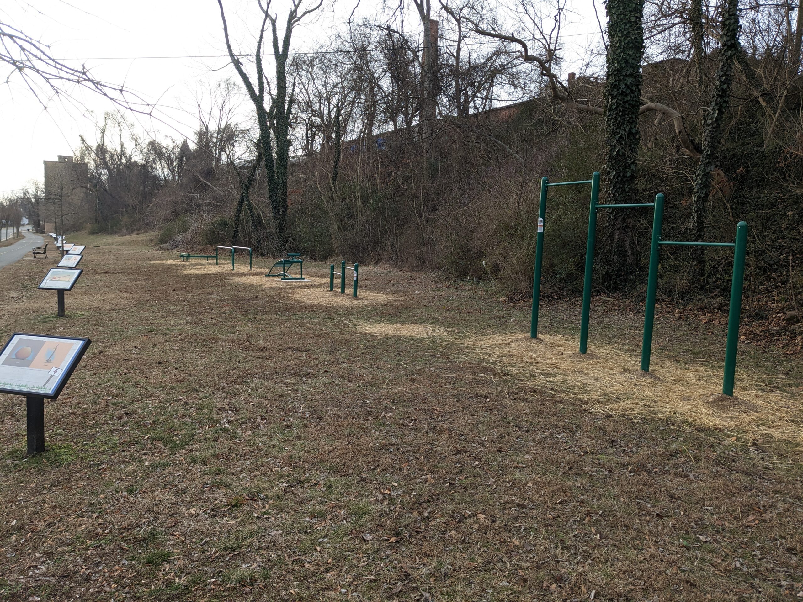 A picture of some green fitness equipment in front of a hillside. The lawn is brown and the trees are bare since it is winter.