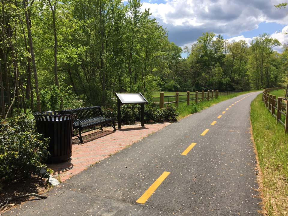 Picture of a bike trail with a rest area along the side with a bench, a trash can, and a historic marker